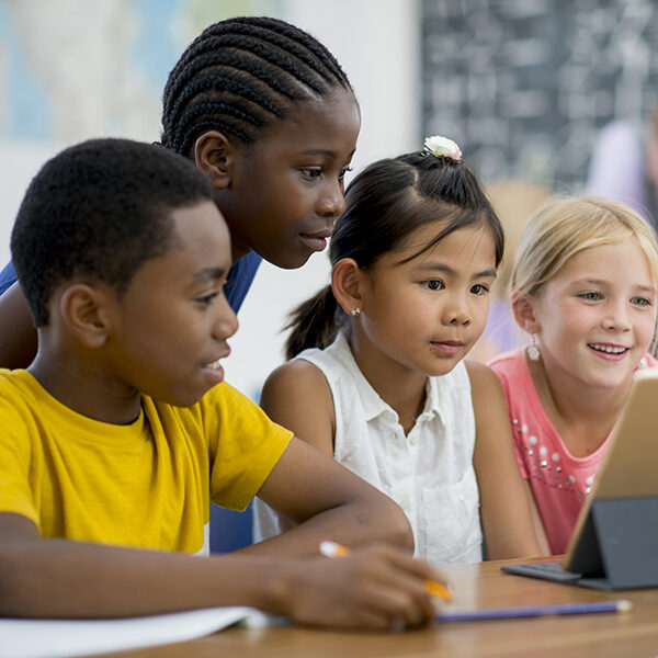 A multi-ethnic group of elementary age children are sitting together and are watching an educational video on a digital tablet.